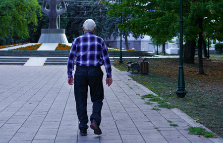An elderly man in a checkered shirt strolls along a pathway in a park during the evening. Surrounding trees provide shade and a peaceful atmosphere as he enjoys his walk.の写真素材