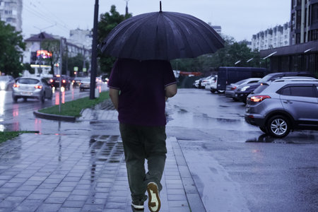 A man walks alone along a rain-soaked city sidewalk, holding a black umbrella. The wet pavement reflects the gray sky, and cars are parked nearby, creating a serene atmosphere.の写真素材