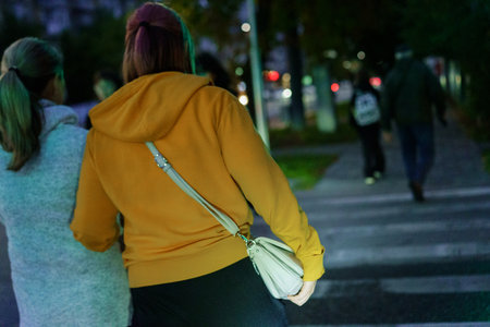 Two women stroll together at night, crossing a city street illuminated by soft streetlights. People...