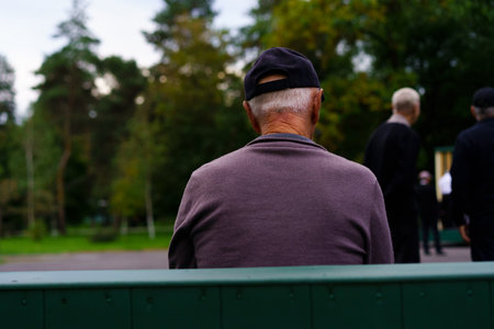 An elderly man is seated on a green bench in a park, wearing a black cap, quietly watching people around him as evening approaches. Trees provide a serene backdrop.の写真素材