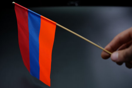 A small Armenian flag featuring blue, red, and orange stripes is being held by a hand. The...