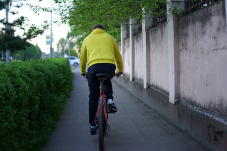 A person wearing a bright yellow hoodie rides a bicycle along a peaceful path lined with lush green bushes and trees during the early evening hours.の写真素材