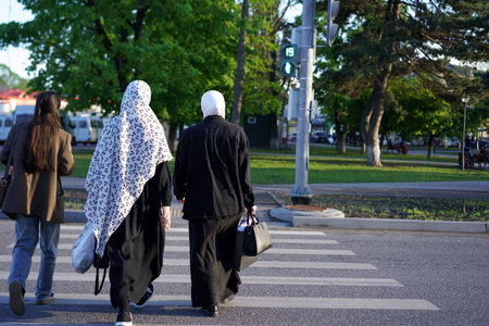 Three women walk together across a zebra crossing, two in traditional attire and one casually dressed, under clear skies near a green area.の写真素材