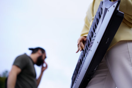 A musician holds a keyboard, focusing on their performance in a park. A friend stands nearby, looking thoughtful as they enjoy the day under a clear sky.の写真素材