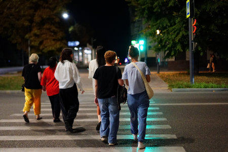A group of six friends walks together on a city street, crossing a crosswalk with traffic lights glowing green. The scene captures the lively atmosphere of a city night.の写真素材