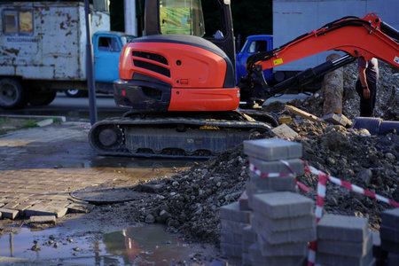 A small excavator is being used at a construction site near a road. The ground is filled with dirt...