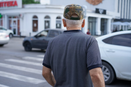 An elderly man with gray hair is walking towards a busy street crossing. He wears a camouflage cap and a polo shirt. Cars are passing by in an urban setting on a sunny day.の写真素材