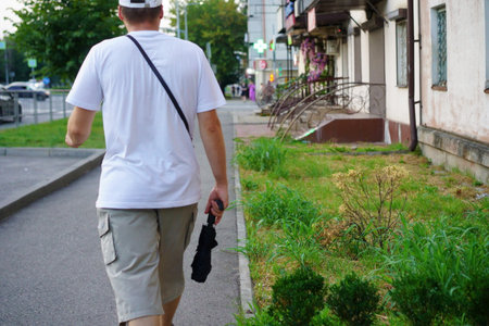 A man in a white shirt and shorts walks along a sidewalk on a sunny afternoon, holding a black umbrella. Green grass and buildings surround him.の写真素材