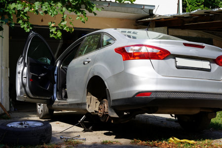 A silver car is elevated on a jack inside a home garage. One wheel is removed, and tools and a tire are nearby. Sunlight filters through the open door, illuminating the workspace.の写真素材