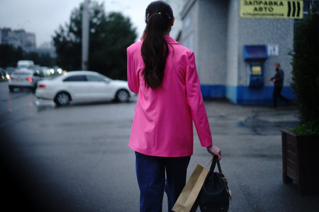 A woman dressed in a bright pink blazer walks along a wet city street. She holds shopping bags in one hand while navigating traffic and pedestrians in the evening.の写真素材