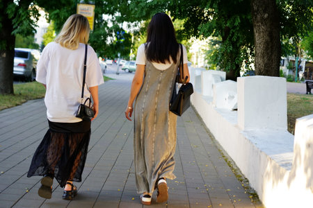 Two women stroll side by side along a sidewalk in a city park, both dressed in summer outfits. They...