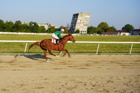 A jockey in green and white rides a chestnut horse at high speed on a sandy race track. The sun shines brightly, illuminating the surrounding area, including distant buildings.の写真素材