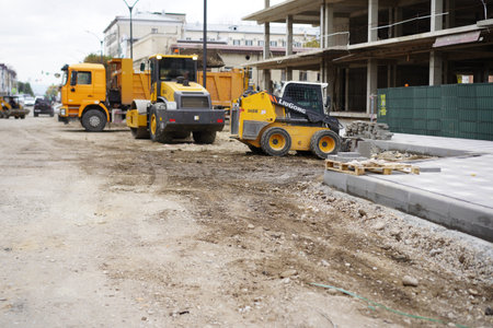 Heavy construction vehicles work on a dirt road in an urban setting. A skid steer loader and dump...
