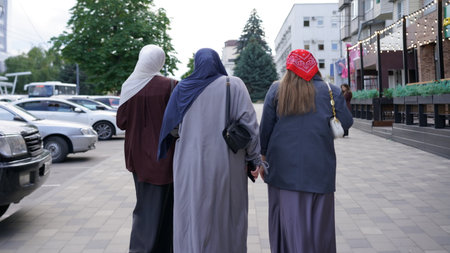 Three women dressed in long garments stroll down a city street in the early evening. Trees line the path, suggesting a lively urban environment with nearby cars.の写真素材