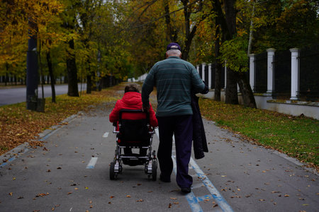 An elderly man walks gently along a paved path, pushing a child in a stroller surrounded by colorful autumn leaves. The atmosphere is calm and peaceful.の写真素材