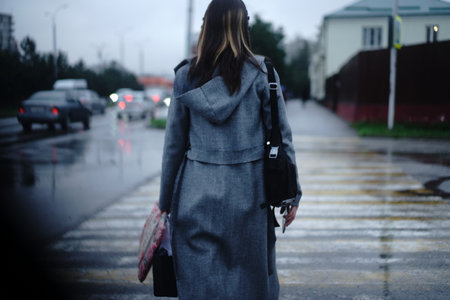 A woman in a gray coat walks through a wet street while holding an umbrella. Rain falls softly as she crosses a crosswalk in a city setting during the evening.の写真素材