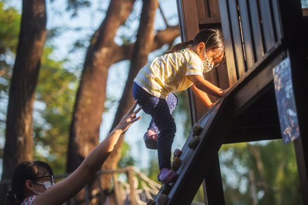 little girl climbs up a wooden wall in the playground with her mother behind her to keep safetyの写真素材