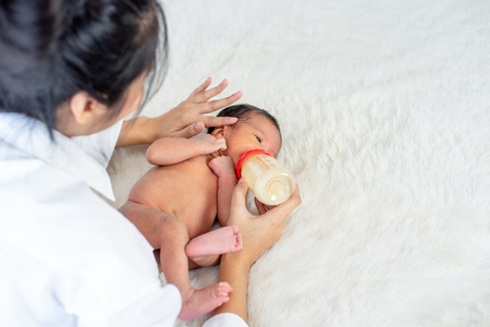 Asian mother give milk feeding with bottle to little newborn baby on white bed and take careの写真素材