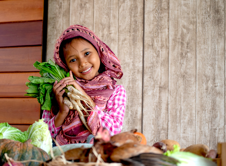 Asian little young girl look forward and smile among various types of vegetable also hold the radish behind hen on the table in her kitchenの写真素材
