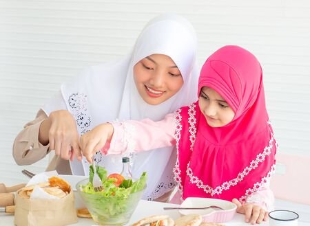 Muslim mother point to vegetable salad while little girl with pink hijab has fun with mixing salad put on the tableの写真素材