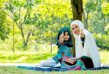 Muslim woman and girl show thumps up to the camera during reading some books in the garden.の写真素材
