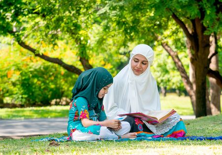 Muslim mother teach her daughter to read religion textbook for understanding the way of good life. They stay in the green garden during day time.の写真素材
