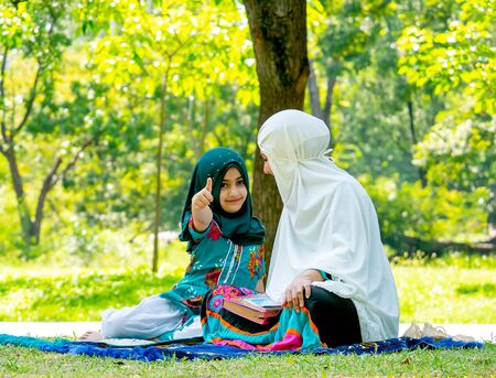 Muslim woman look at her child and girl show thumps up to the camera during reading some books in the garden.の写真素材