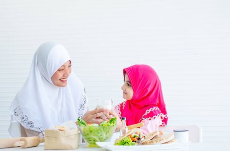 Muslim mother serve glass of milk to her little girl and also look to each other with smiling, bowl of vegetable salad on the table in front of white background.の写真素材