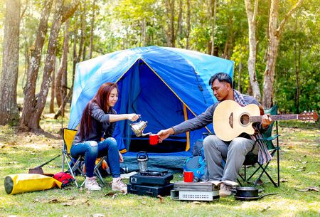 Couple of lover are camping near the forest have relaxing activity with man play guitar and woman give him with a cup of coffee.の写真素材
