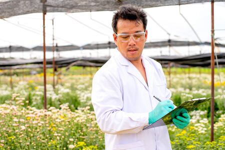Asian scientist man with white lab gown analysis and record the data of white and multicolor flowers in the garden during day time.の写真素材