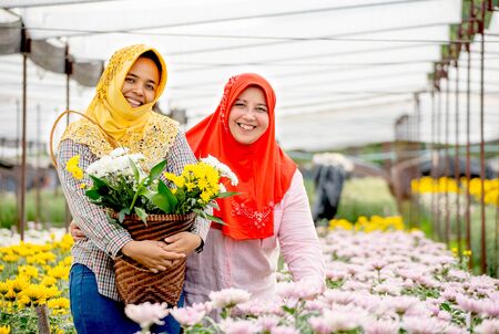 Two Muslim worker girls are standing and smiling among pink and yellow flowers in the garden during day time. One wear yellow hijab and the other one is red hijab.の写真素材