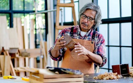Old Asian craftsman hold wooden small house and look to it, in the room with wood pile and surround with glasses.の写真素材