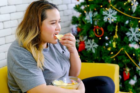 Fat girl is eating potatoes chips and watching TV in her house during Christmas time with concept relaxation at home during Christmas holiday.の写真素材