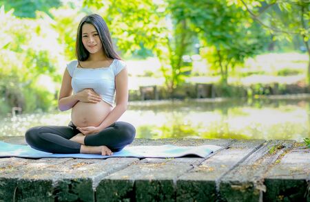 Beautiful Asian pregnant woman look forward while sit on mat on the wood bridge and also touch her belly with morning light and look relaxation.の写真素材