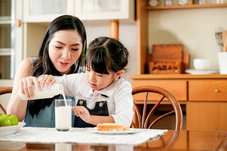 Asian mother take care her daughter pour milk to a glass on the table in house kitchen during breakfast meal. Main focus is the mother.の写真素材
