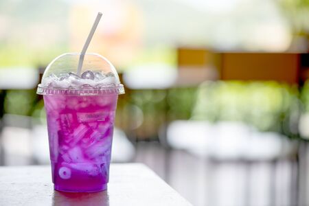 Purple lemonade drink in plastic glass and tube are put on the table with day light and coffee shop as background.の写真素材