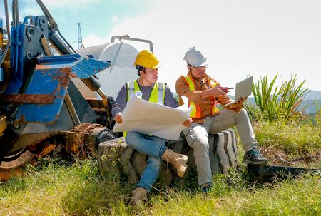 Engineer manager with laptop discuss with his team and sit on big wheel of tractor.