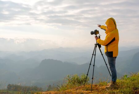 The photographer with yellow jacket stand near to his camera and stand in front cliff, cloudy and...
