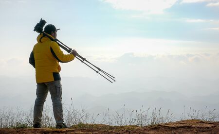 The photographer carry camera with tripod on his shoulder and look to the mountain over the cliff with morning light.の写真素材