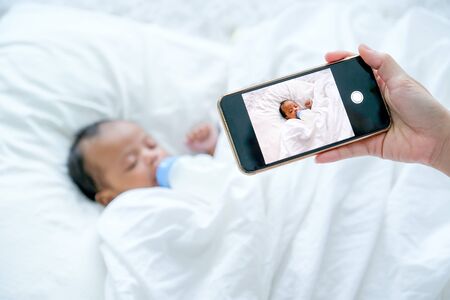 Mother hand hold mobile phone to take a photo of newborn baby during she is sleeping on white bed.の写真素材