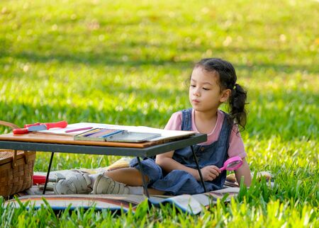 Cute Asian little girl hold magnifier glass and sit near table with colored pencils and toys in the garden with morning light.の写真素材