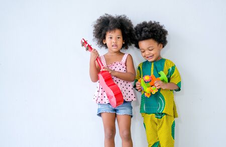 Two young African brother and sister  stand in front of white wall and girl holds small guitar and boy holds some toys in hand with day light.の写真素材