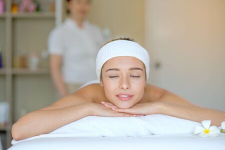 Portrait of White beautiful woman lie on bed in spa room and look relax and happy with Thai masseuse stand behind as background.の写真素材