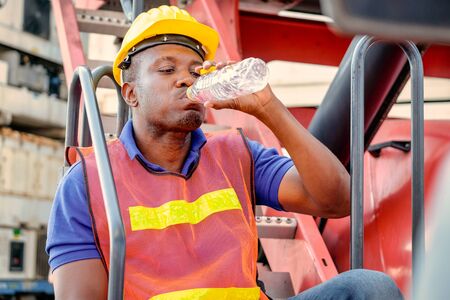 Portrait of African worker man drink water from bottle and stay on big crane truck in cargo shipping area.の写真素材
