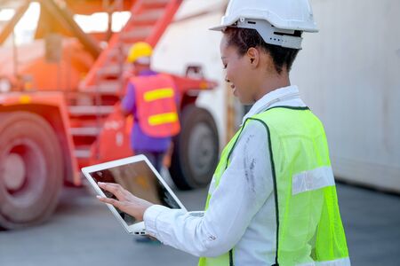 African technician worker woman touch and look to laptop while her co-worker work with construction vehicle.の写真素材