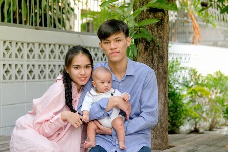 Asian mother is sitting with her husband who is holding their little baby in home garden with morning light and they look happy.の写真素材
