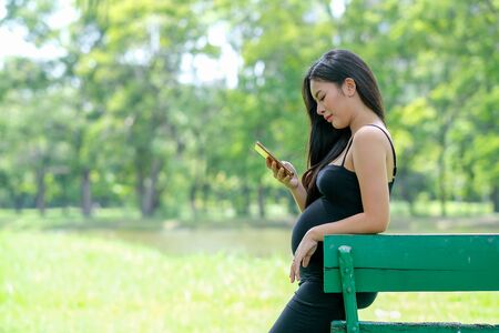 Portrait of beautiful pregnant woman with black dress use mobile phone in park or garden with day light and she look relax.の写真素材