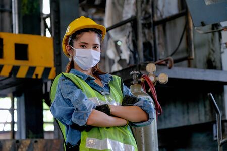 Factory woman worker or technician with hygienic mask stand with confident action with big machine as background.の写真素材