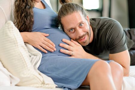 Man sit and listen baby sound from his pregnancy wife belly and he look happy also look to camera. Concept of good relationship of family member support good quality of life in new normal lifestyle.の写真素材