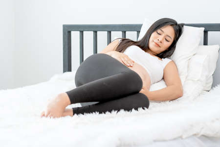Beautiful Asian pregnant woman hold glass of milk and look outside through white curtain and she...